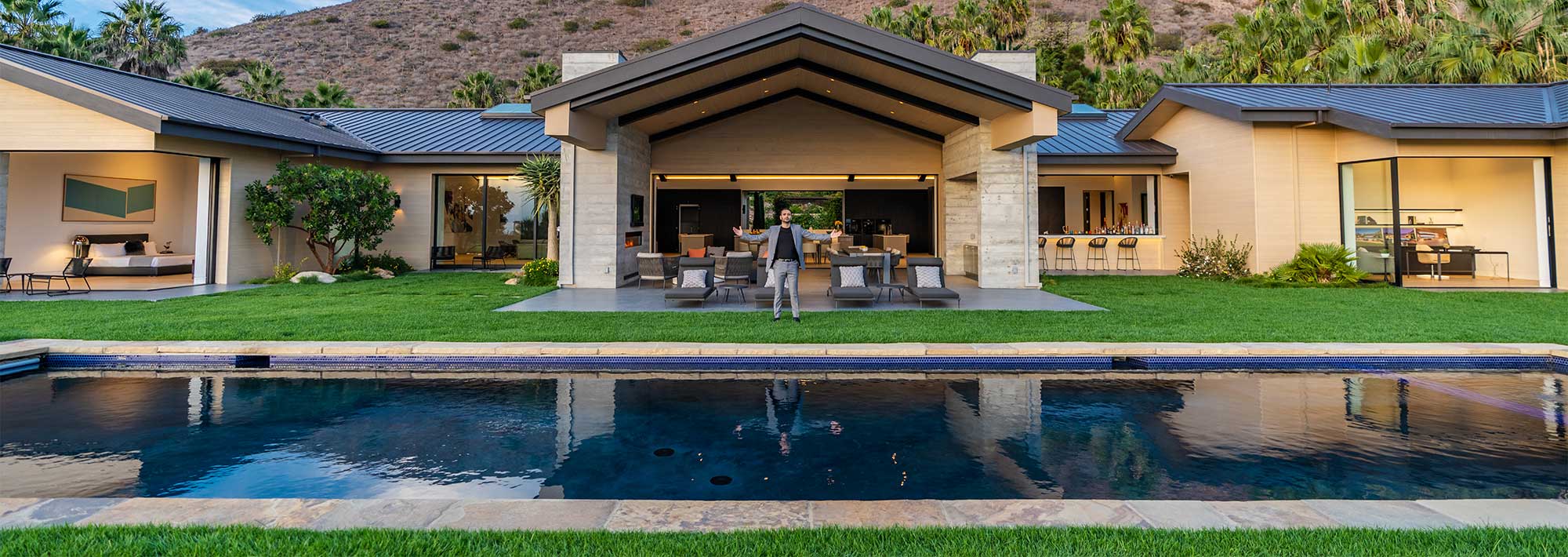 Modern hillside villa with solar panels, a central pool, and surrounding palm trees. Open-air design with expansive glass walls and green lawn. Desert landscape in the background.